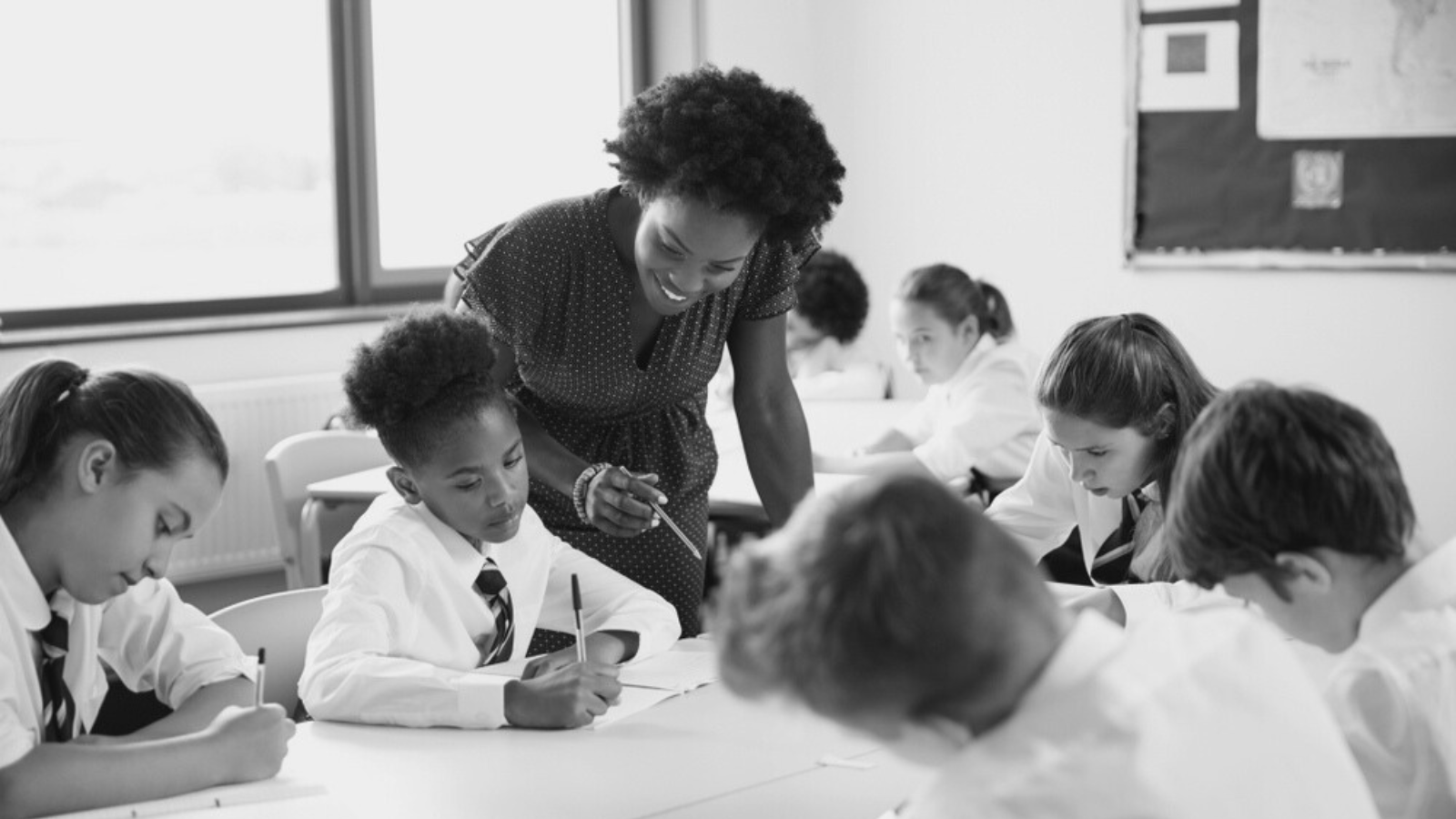 A black-and-white picture shows a smiling teacher leaning over to assist a student in a bright, focused classroom where children in uniform are diligently working at their desks