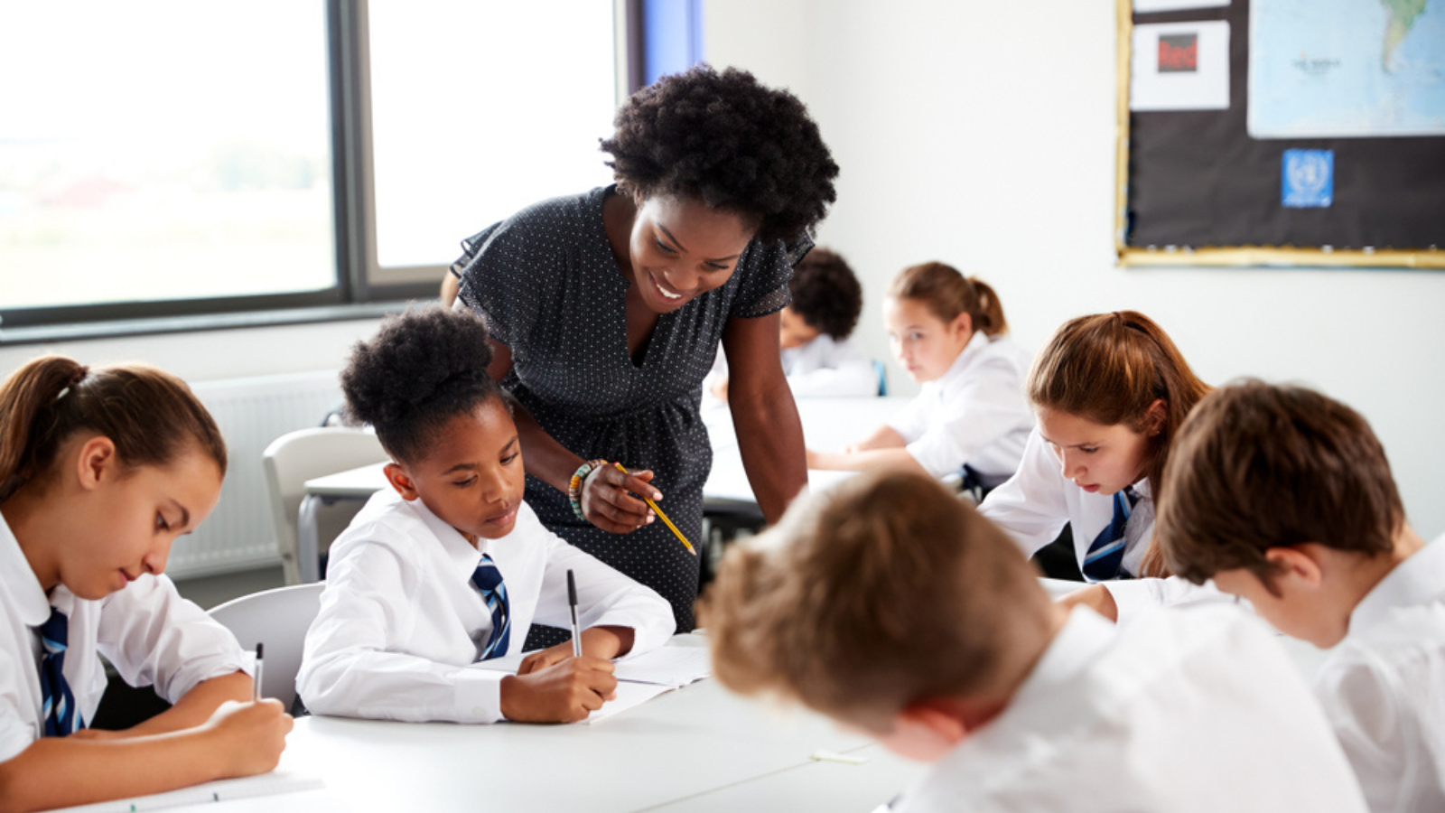A coloured saturated picture shows a smiling teacher leaning over to assist a student in a bright, focused classroom where children in uniform are diligently working at their desks